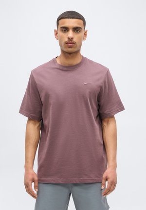 Νέος άνδρας με κοντά σκούρα μαλλιά που φοράει μωβ μπλουζάκι Nike και ανοιχτό γκρι σορτς, στέκεται μπροστά σε απλό λευκό φόντο.