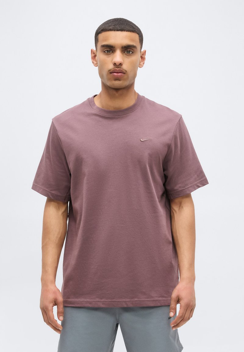 Mladý muž s krátkymi tmavými vlasmi v tričku Nike mauve farby a svetlosivých kraťasoch stojaci pred bielym jednoliatym pozadím.
