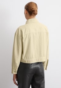 Light yellow cropped jacket with a collar and long sleeves, made of a smooth fabric; back features a horizontal seam. Worn with black pants.