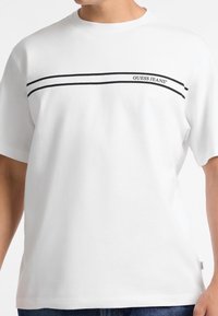 Bijela pamučna majica kratkih rukava, s crnim horizontalnim prugama i logom "GUESS JEANS" centriranim iznad pruga.