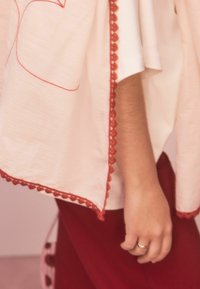 Hellrosa Kleidungsstück mit roten, gewellten Abschlüssen, getragen über einem weißen Oberteil, kombiniert mit einem dunkelroten Unterteil. Hand mit einem Ring sichtbar.