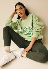 Lysgrøn sweatshirt med "Montreal" tekst, parret med mørkegrønne leggings og hvide sneakers. Tan sokker fremhæver outfittet. Glatte teksturer.