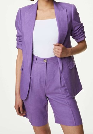 Kvinde iført en skræddersyet lilla blazer med rynkede ærmer og matchende højtaljede shorts, kombineret med en hvid tætsiddende top.