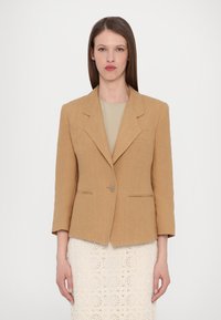 Lys brun lin blazer med skreddersydd passform, klaffelapper, enkelt knappelukking og to frontlommer. Brukt over en beige topp.