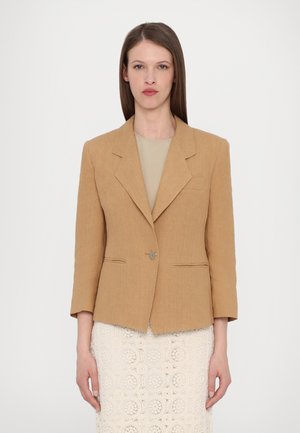 Lys brun lin blazer med skreddersydd passform, klaffelapper, enkelt knappelukking og to frontlommer. Brukt over en beige topp.