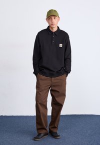 Polo shirt nero a maniche lunghe con tre bottoni e una piccola patch con logo, abbinato a pantaloni marroni e sneakers nere. Cappellino verde oliva.