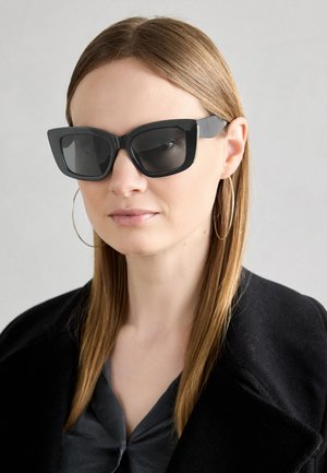 Sorte katteøyne solbriller med mørke linser, tykke rammer og subtile detaljer. Båret med en svart frakk og grå topp, med langt, rett hår.