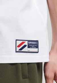 T-shirt bianco in cotone con un'etichetta blu, rossa e bianca in fondo che presenta "SUPERDRY" e dettagli sulla posizione. Pantaloni verde oliva visibili.