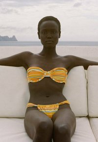 Bandeau bikini z rumenimi črtami s ukrivljenim detajlom; ujemajoče se nizko pasovno dno, gladka tekstura in priljubljenost. Nevtralna ozadje.