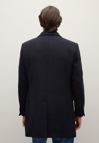 Cappotto nero dal design sartoriale, tessuto testurizzato, rever a incavo, spacco singolo e polsini con bottoni. Indossato sopra jeans blu. Vista posteriore.