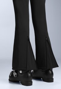 Dolne nogi w czarnych spodniach z tylnymi zamkami, rozszerzających się nad czarnymi butami narciarskimi z srebnymi klamrami.