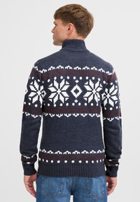 Gebreide coltrui in marineblauw met een wit en rood sneeuwvlokpatroon. Heeft ribgebreide manchetten en zoom, en een gestructureerde afwerking.