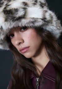 Giovane donna con lunghi capelli scuri che indossa un cappello in pelliccia sintetica bianco e grigio e una giacca a zip viola scuro, che guarda leggermente di lato.