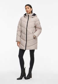 Ljusbeige pufferjacka med huva, dragkedja framtill och sidofickor. Har horisontellt sydda paneler och svarta dragkedjespännen.
