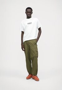 Weißes T-Shirt mit gedrucktem Logo, olivgrüne Cargo-Hosen mit Taschen und orangefarbenes Schuhwerk mit Schnürsenkeln und Gummisohlen.