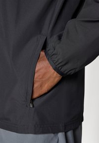 Black lightweight jacket with elastic cuffs, zippered side pocket, and smooth texture. Features a straight hemline and minimalist design.