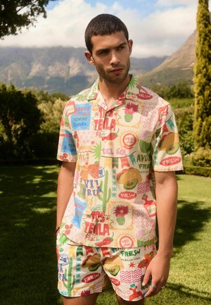 Muž v pestrofarebnej krátkej košeli a zodpovedajúcich šortkách s motívom mexického jedla stojí vonku s horami a zeleňou v pozadí.