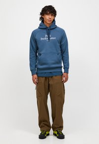 Blauwe hoodie met de tekst "Peak Performance", kangoeroezak, en geribbelde manchetten. In combinatie met een bruine cargobroek en zwarte schoenen met groene accenten.