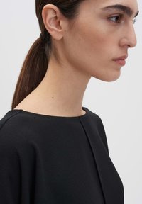 Indumento nero con finitura testurizzata, caratterizzato da un ampio scollo a barca e una forma dritta, mostrato di profilo con i capelli raccolti in una coda di cavallo.