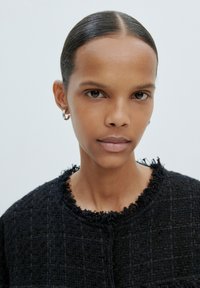 Zwarte textuurjasje met rafelige halslijn, met een geweven patroon. Model heeft steil haar en subtiele make-up, tegen een lichte achtergrond.
