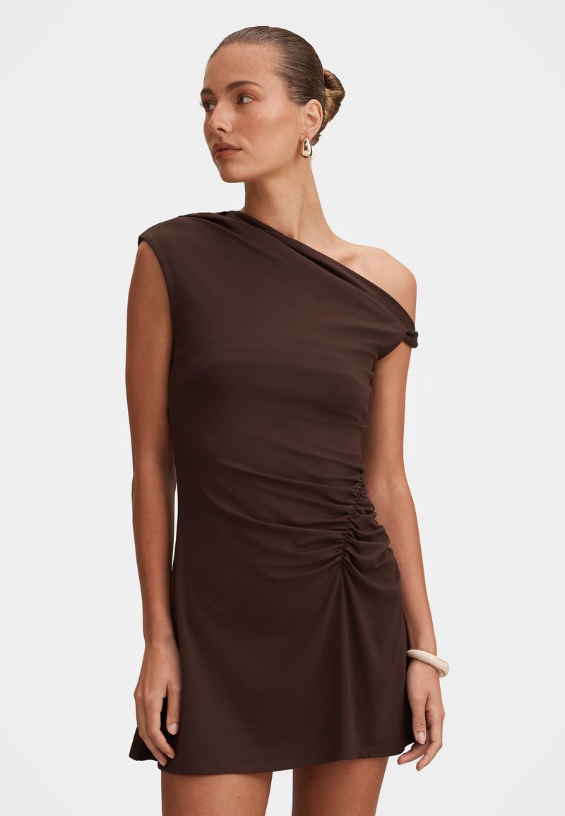 Γυναίκα που φοράει ένα μίνι φόρεμα μονό ώμου σε σκούρο καφέ χρώμα με ραφές στη μία πλευρά, τα μαλλιά της είναι δεμένα κότσος, φοράει χρυσούς κρίκους στα αυτιά και ένα λευκό βραχιόλι.