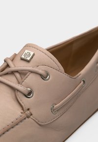 Mocasini bej din piele cu șireturi încrucișate, cu găuri metalice și un mic logo pătrat metalic pe partea superioară din față.