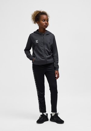 Junge Frau steht mit einer Hand in der Tasche, trägt einen schwarzen Hoodie mit Reißverschluss und schwarze Jogginghose mit Chevron-Streifen an den Seiten.