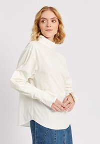 Blondide juustega naine, kes kannab loose-stiilis valget pikkade varrukatega toppi ja sinist teksaseelikut, vaatab külje peale, käed kokku pandud.