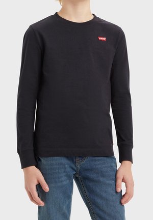 Dziecko ubrane w czarną koszulkę Levi's z długim rękawem z czerwonym logo na piersi oraz niebieskie dżinsy, stojące na jednolitym jasnym tle.