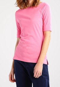 Person med en rosa kortärmad topp och mörkblå byxor, som står mot en enkel vit bakgrund.