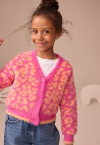 Lyserød og gul fluffy cardigan med leopardmønster, der har V-hals og tre lyserøde knapper. Kombineret med blå jeans.