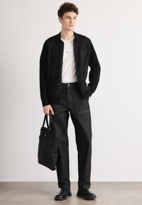 Črni bombažni jopič nad belim majico, v kombinaciji s temnimi jeans hlačami. Model drži črno torbo in nosi črne čevlje.