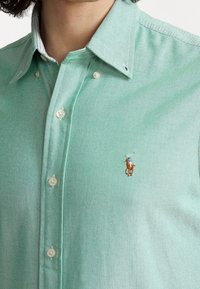 Grön skjorta med knappar i texturerat tyg, med en polospelarlogotyp på vänster bröst och med en kontrasterande ljusfärgad krage.