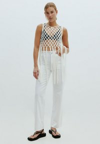 Witte gebreide crop top met een geometrisch patroon en franjes, gecombineerd met witte rechte broek en zwarte teenslippers.