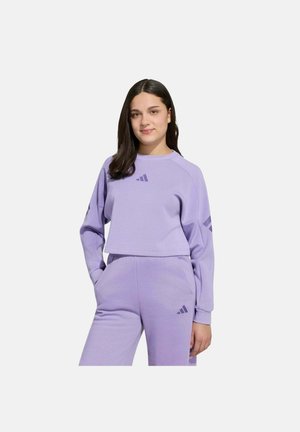 Jeune femme aux cheveux foncés portant un sweat-shirt violet clair et un pantalon assorti avec des logos Adidas, debout, les mains dans les poches.