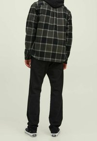 Camicia in flanella a quadri neri e grigi con maniche lunghe, abbinata a pantaloni neri e sneakers neri, vista da dietro su uno sfondo chiaro.