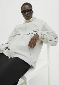 Lys beige zip-up jakke med sorte detaljer og elastiske manchetter, parret med sorte bukser. Modellen bærer sorte solbriller. Minimalistisk design.