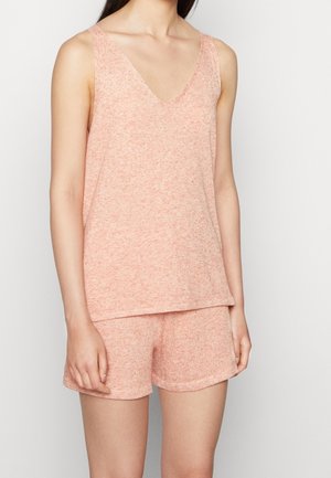 Femme portant un haut sans manches rose clair avec col en V et un short assorti, debout devant un fond blanc uni.