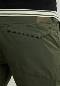 Primer plano de pantalones verde oscuro con un bolsillo trasero y una etiqueta marrón que dice "CHASIN'" encima del bolsillo, combinados con una camisa de rayas.