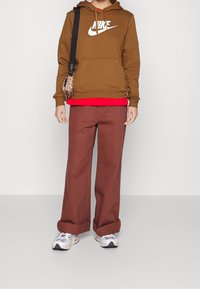 Brun Nike huvtröja, röd T-shirt under, bruna vida byxor och grå-vita sneakers. Liten leopardmönstrad väska på axeln.