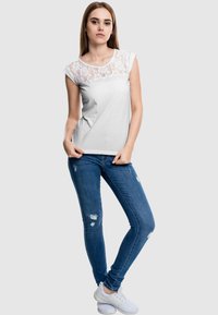 Camiseta blanca con panel de encaje con patrón de rosas en los hombros, combinada con jeans azules rasgados y zapatillas blancas. Diseño casual y ajustado.