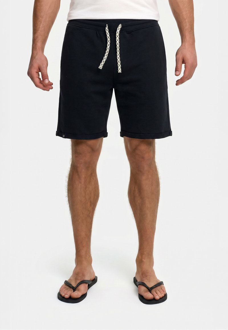 Homme portant un short noir à cordon, un t-shirt blanc et des tongs noires, debout, les jambes légèrement écartées sur un fond uni.