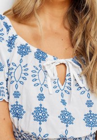 Blouse blanche avec broderie florale bleue, design décolleté, devant en trou de serrure avec fermeture à lacer, et manches volantes.