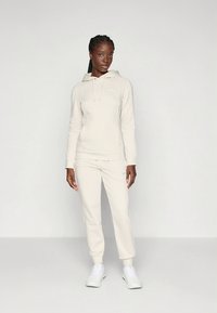 Világos bézs kapucnis pulóver elülső kenguruzsebbel és hozzáillő melegítőnadrággal. Puha anyagból készült, logó részlettel.