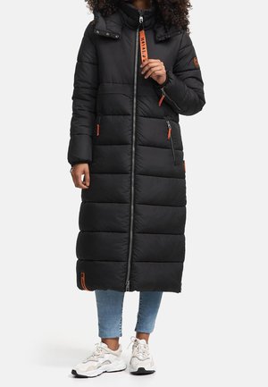 Personne portant un long manteau noir matelassé avec des tirettes de fermeture éclair orange, un jean court bleu et des baskets blanches sur un fond uni.