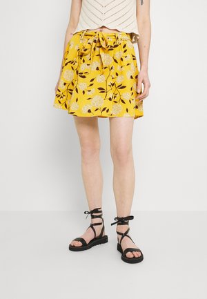 Femme portant une jupe fleurie jaune avec une ceinture nouée à la taille et des sandales plates noires à brides, debout devant un fond clair uni.
