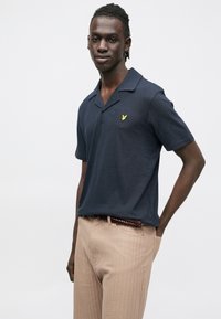 Tengerészkék rövid ujjú póló lapos gallérral, melyen egy kis sárga logó található a mellrészen; bézs, texturált nadrággal párosítva.