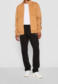 Giacca beige con cerniera e pannelli laterali, t-shirt bianca sotto, pantaloni della tuta neri e sneakers bianche. Design semplice e stile casual.