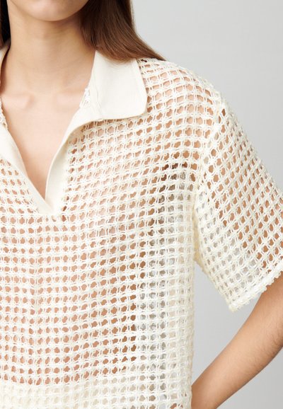 Chemise à col et manches courtes en crème avec un design en maille ouverte. Présente une texture légère et une coupe ample, idéale pour les superpositions.