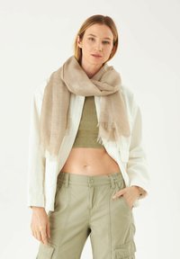 Foulard beige léger avec des bords frangés, drapé sur un mannequin portant un top olivier court et une chemise blanche, associé à un pantalon cargo.